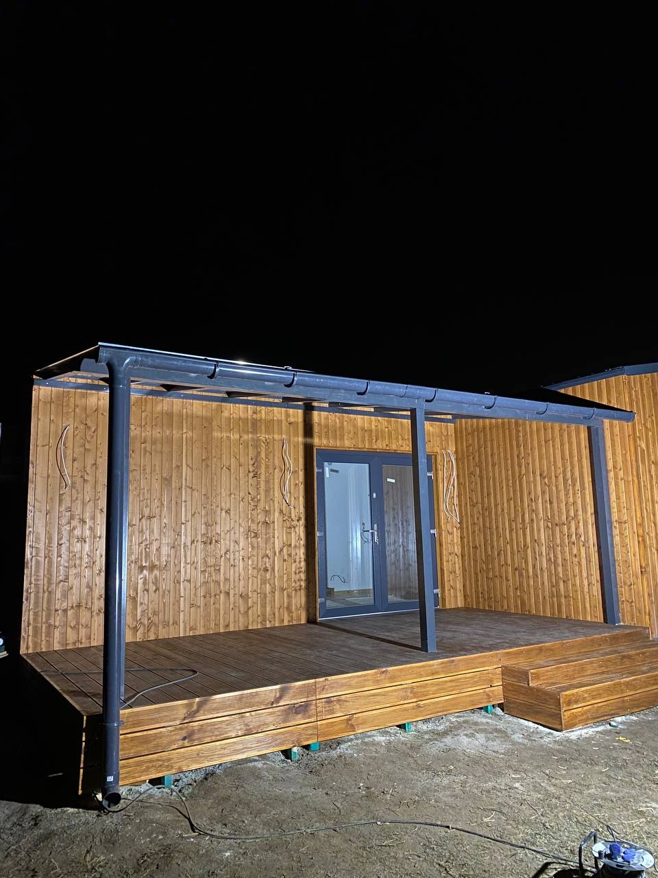 Drewniany taras z zadaszeniem z metalową konstrukcją, oświetlony nocą, widoczne schody i drzwi wejściowe do budynku.