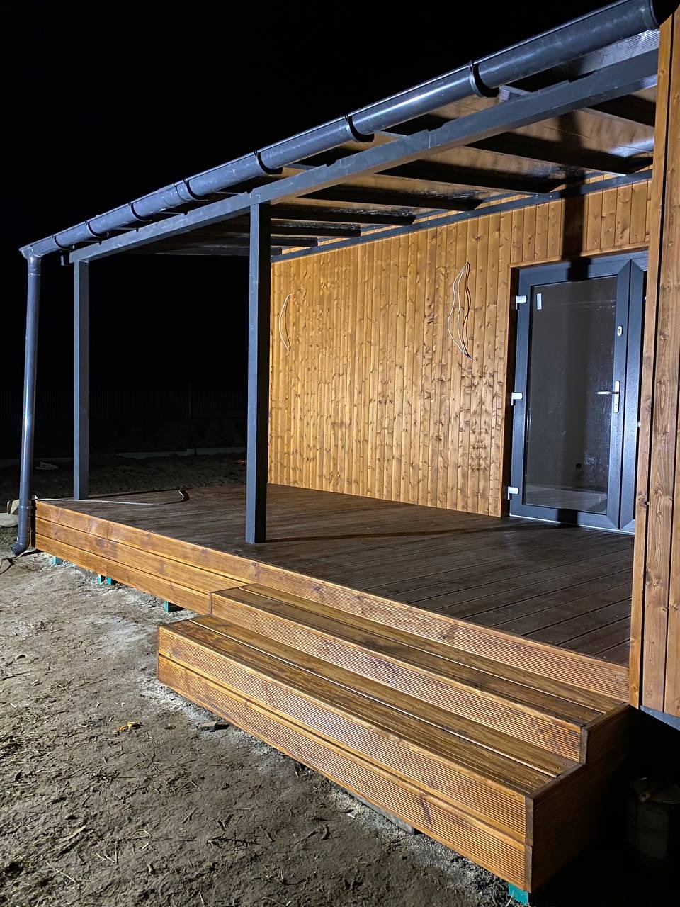 Nowoczesny taras z drewna z zadaszeniem metalowym, oświetlony w nocy, z widocznymi schodami wejściowymi i drzwiami do budynku.