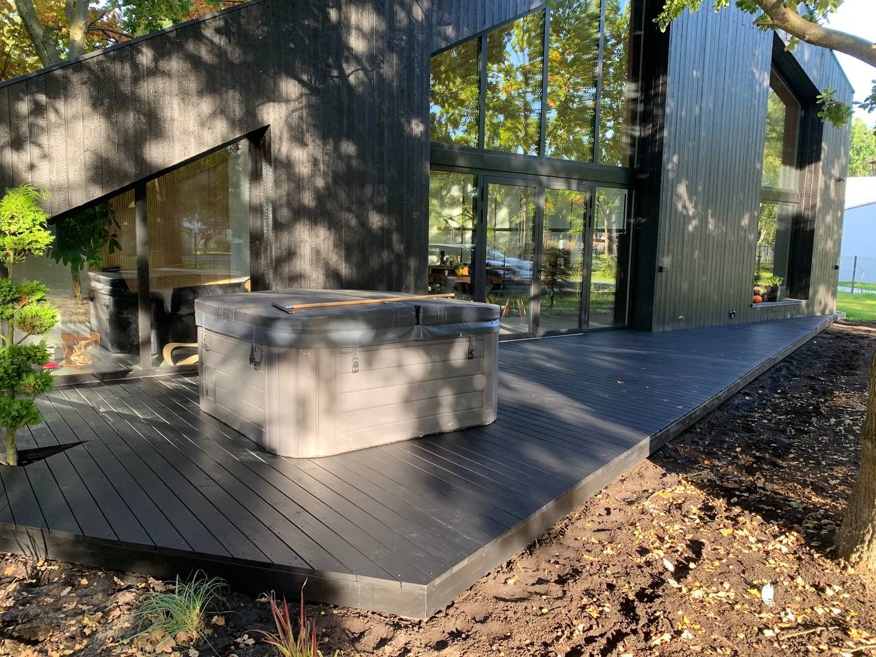 Nowoczesny dom z czarną fasadą i dużymi oknami, zadaszony taras z ciemnego drewna z jacuzzi, widoczne świeżo obsadzone rośliny w ziemi.