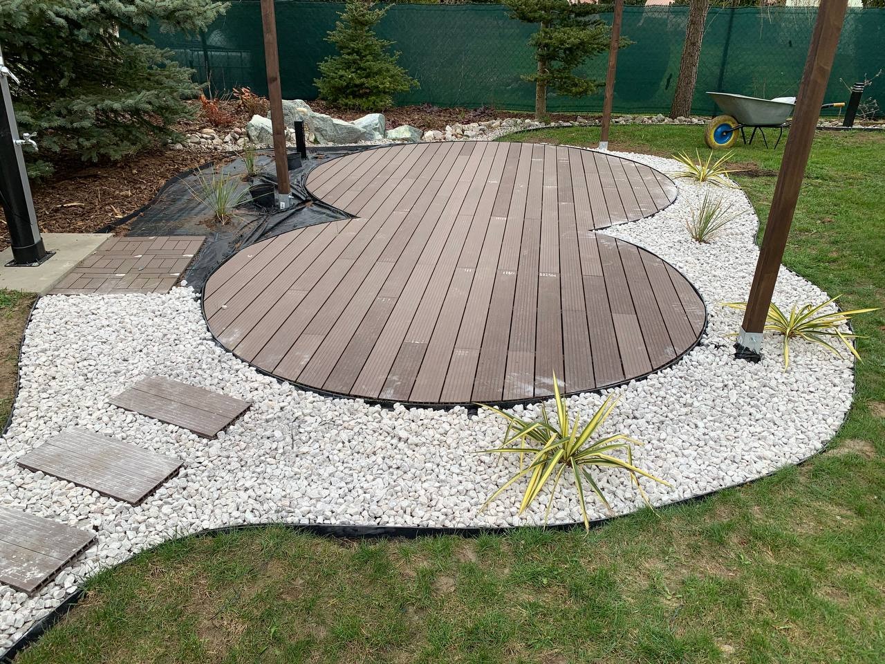 Kreatywnie ukształtowany, brązowy podest z desek kompozytowych otoczony białymi kamykami i roślinami ozdobnymi, z drewnianymi słupami i ścieżką z płyt chodnikowych w ogrodzie.