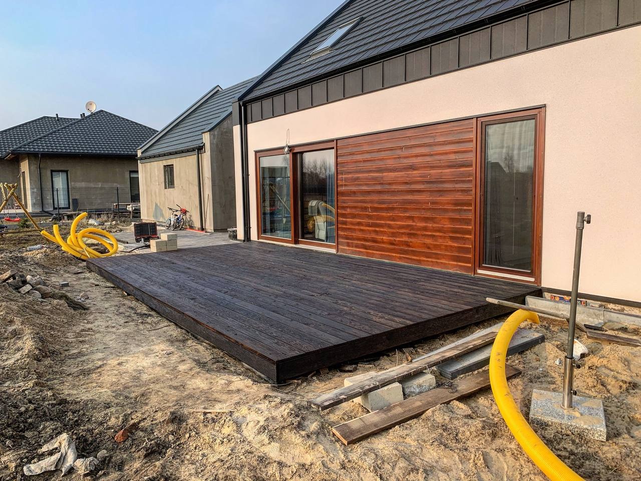 Ciemny, drewniany taras przylegający do domu z drewnianą elewacją, widoczne żółte węże i materiały budowlane sugerujące trwające prace wykończeniowe.