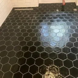 Błyszcząca, czarna podłoga w łazience wyłożona heksagonalnymi płytkami z jasną fugą, widoczny odpływ liniowy i fragment białej ściany z płytek.