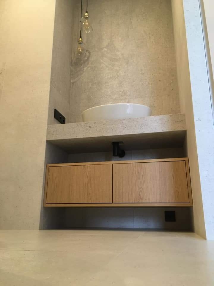 Minimalistyczna łazienka z umywalką nablatową, drewnianą szafką pod umywalką i trzema wiszącymi żarówkami na tle ściany wykończonej płytami imitującymi beton.