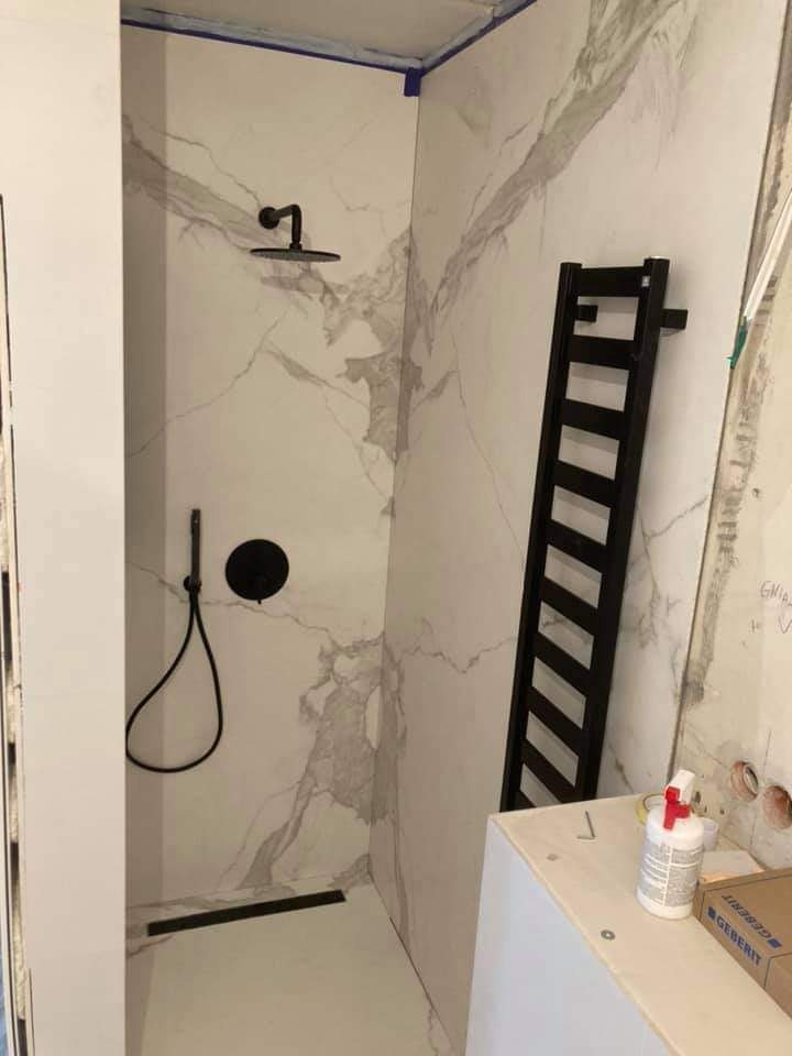 Nowoczesna kabina prysznicowa z marmurowymi płytkami, czarną armaturą i grzejnikiem drabinkowym w trakcie montażu, widoczne materiały budowlane.
