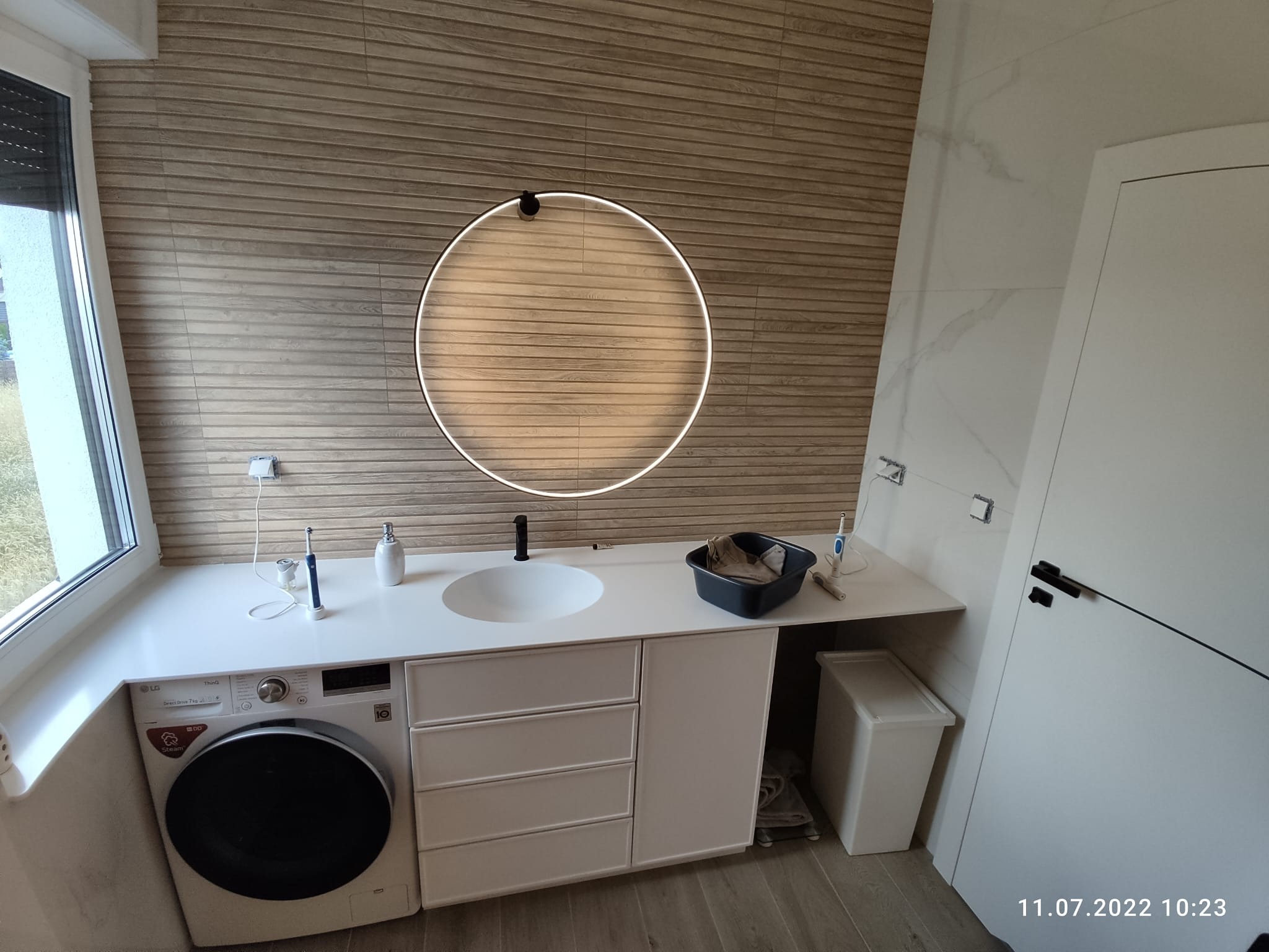 Nowoczesna łazienka w Poznaniu z drewnopodobnymi płytkami na ścianie, okrągłym lustrem z podświetleniem LED, białą umywalką nablatową i pralką pod blatem.