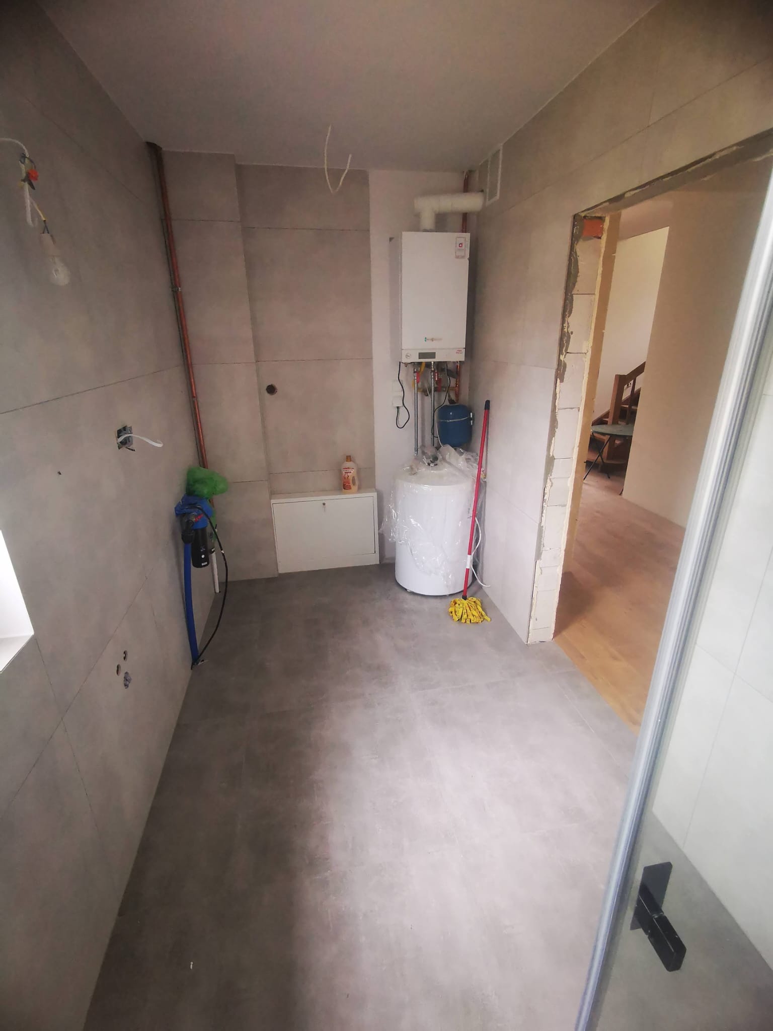 Wnętrze łazienki w trakcie prac wykończeniowych: szare płytki na ścianach i podłodze, bojler owinięty folią, widoczne rury, niezamaskowane otwory w ścianie i fragment futryny drzwi do innego...