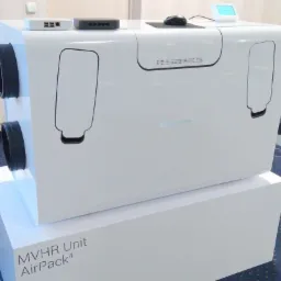 Biała centrala wentylacyjna MVHR Unit AirPack4 z widocznymi otworami wentylacyjnymi, umieszczona na białym postumencie, z routerami i myszką komputerową na górze.