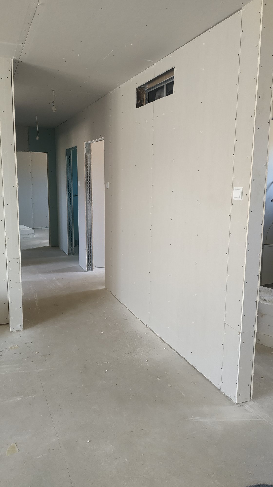 Wnętrze w trakcie montażu płyt gipsowo-kartonowych, widoczny otwór w ścianie przygotowany pod instalację, surowy beton na podłodze i wystające kable elektryczne.