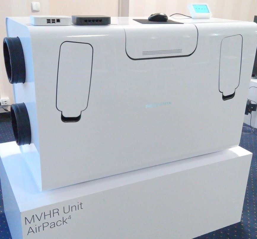 Biała centrala wentylacyjna MVHR Unit AirPack4 z widocznymi otworami wentylacyjnymi, umieszczona na białym postumencie, z routerami i myszką komputerową na górze.