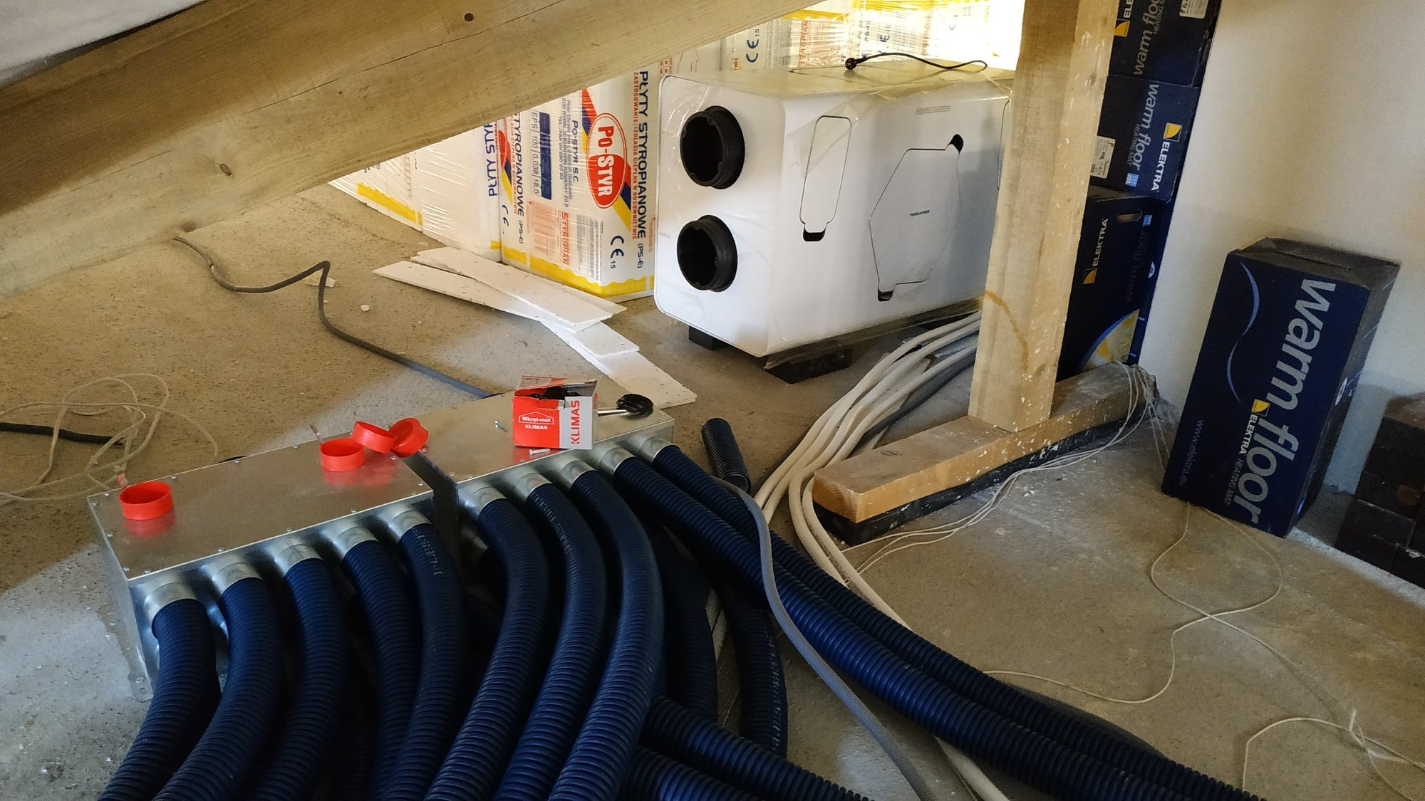 Rozdzielacz wentylacyjny z licznymi niebieskimi przewodami, centrala rekuperacyjna w folii ochronnej, narzędzia i materiały budowlane w surowym wnętrzu poddasza.