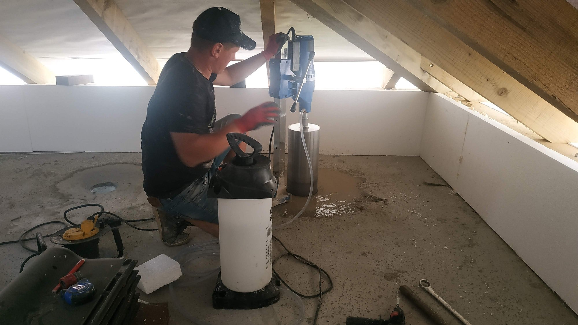 Mężczyzna w czapce i rękawicach roboczych wierci otwór w podłodze na poddaszu za pomocą wiertnicy diamentowej, obok leży wąż z wodą i spryskiwacz ciśnieniowy, a w tle widać białe ściany z izolacji...