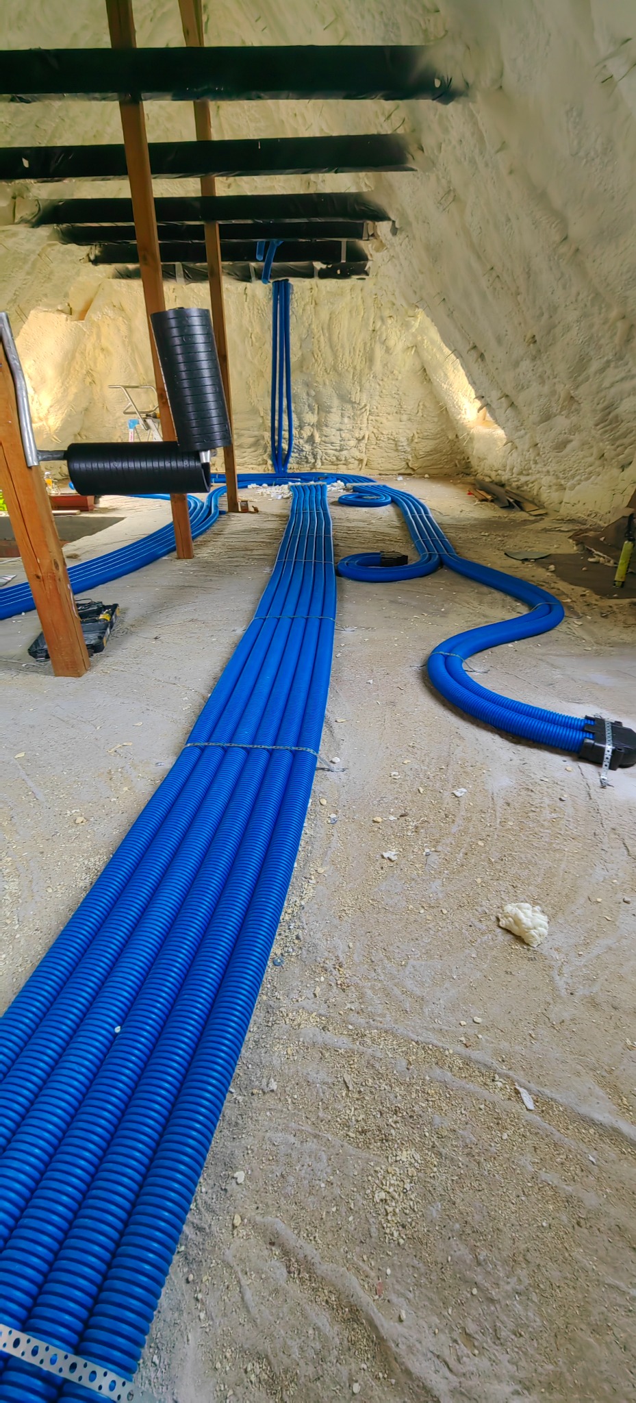 Montaż rekuperacji w poddaszu: wiązka niebieskich, elastycznych przewodów wentylacyjnych ułożona na podłodze, z widoczną izolacją natryskową na ścianach i stropie.