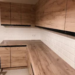 Nowoczesna zabudowa kuchenna z drewnopodobnymi frontami, uzupełniona białymi, prostokątnymi płytkami na ścianie i czarnymi uchwytami.