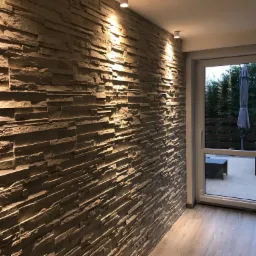 Ściana wyłożona dekoracyjnym kamieniem w odcieniach szarości, oświetlona dwoma punktowymi lampami sufitowymi, z widokiem na taras przez przeszklone drzwi balkonowe.