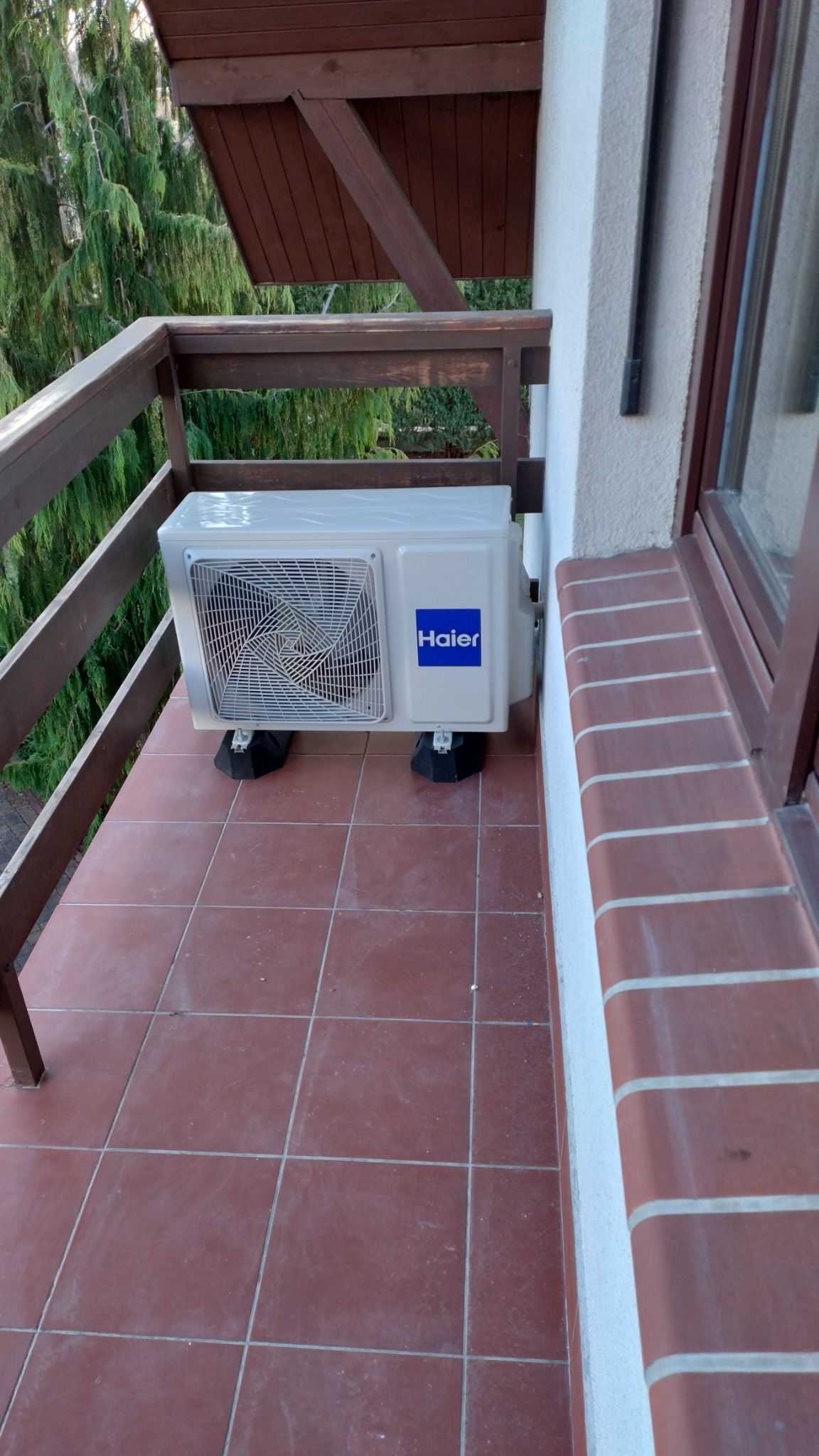 Biała jednostka zewnętrzna klimatyzacji Haier zamontowana na brązowym balkonie z drewnianą balustradą, widok z góry.