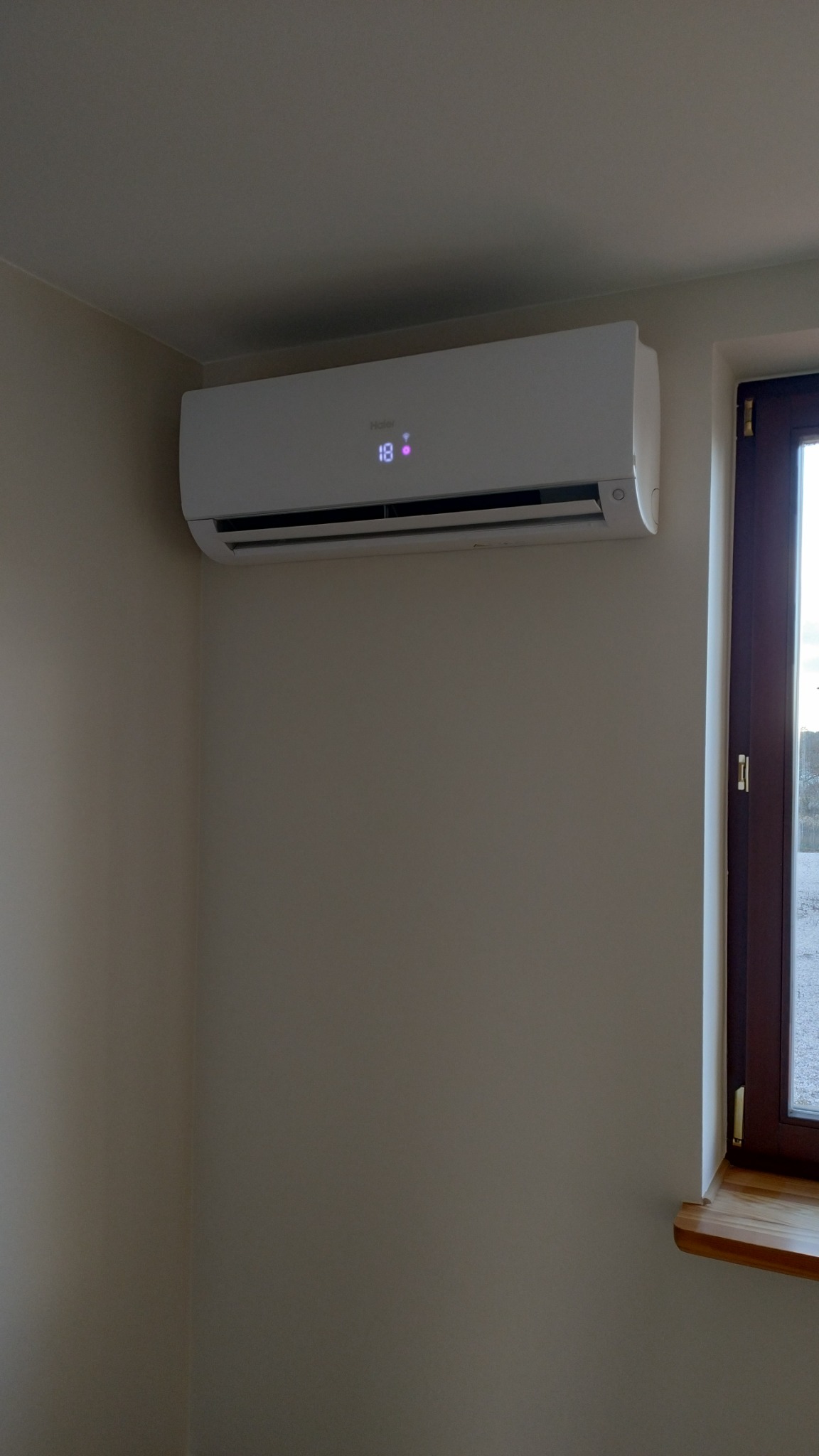 Biały klimatyzator Haier zamontowany na ścianie w jasnym pokoju, obok okna z drewnianą ramą i parapetem.