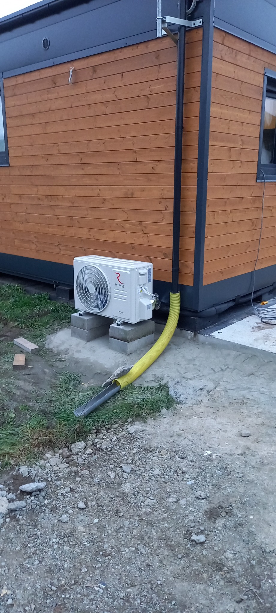 Zewnętrzna jednostka klimatyzacji Rotenso Revo zamontowana na betonowych bloczkach przy drewnianej elewacji budynku, z widoczną żółtą rurą odprowadzającą skropliny.