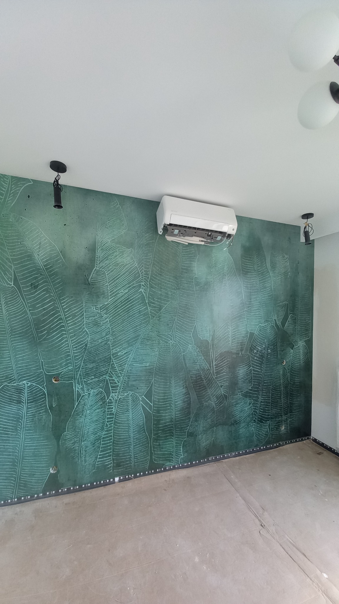 Wnętrze pokoju z dekoracyjną ścianą w odcieniach zieleni z motywem liści, na której zamontowano klimatyzator ścienny w trakcie instalacji, widoczne otwarte elementy urządzenia.