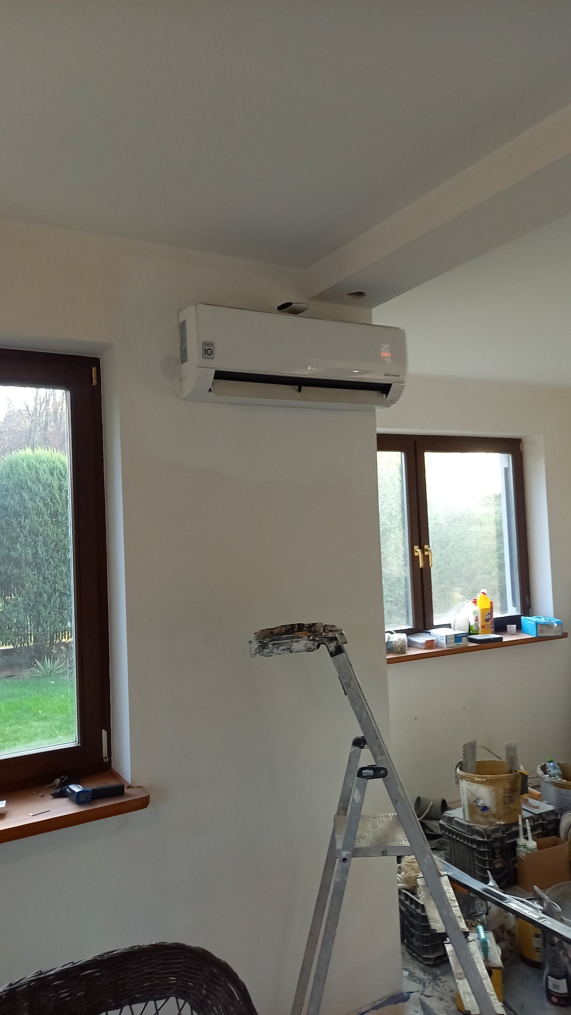 Biały klimatyzator LG zamontowany na ścianie w pokoju, widoczna drabina malarska, okna z widokiem na zieleń i narzędzia na parapecie.