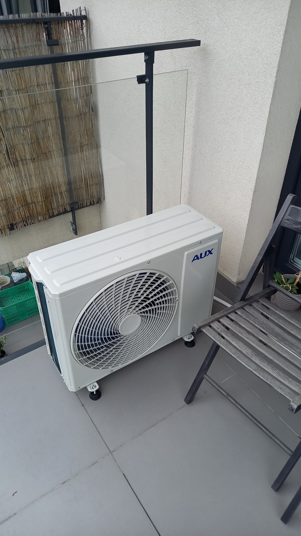 Biała jednostka zewnętrzna klimatyzacji marki AUX umieszczona na szarym balkonie, obok składanego krzesła i szklanej balustrady.