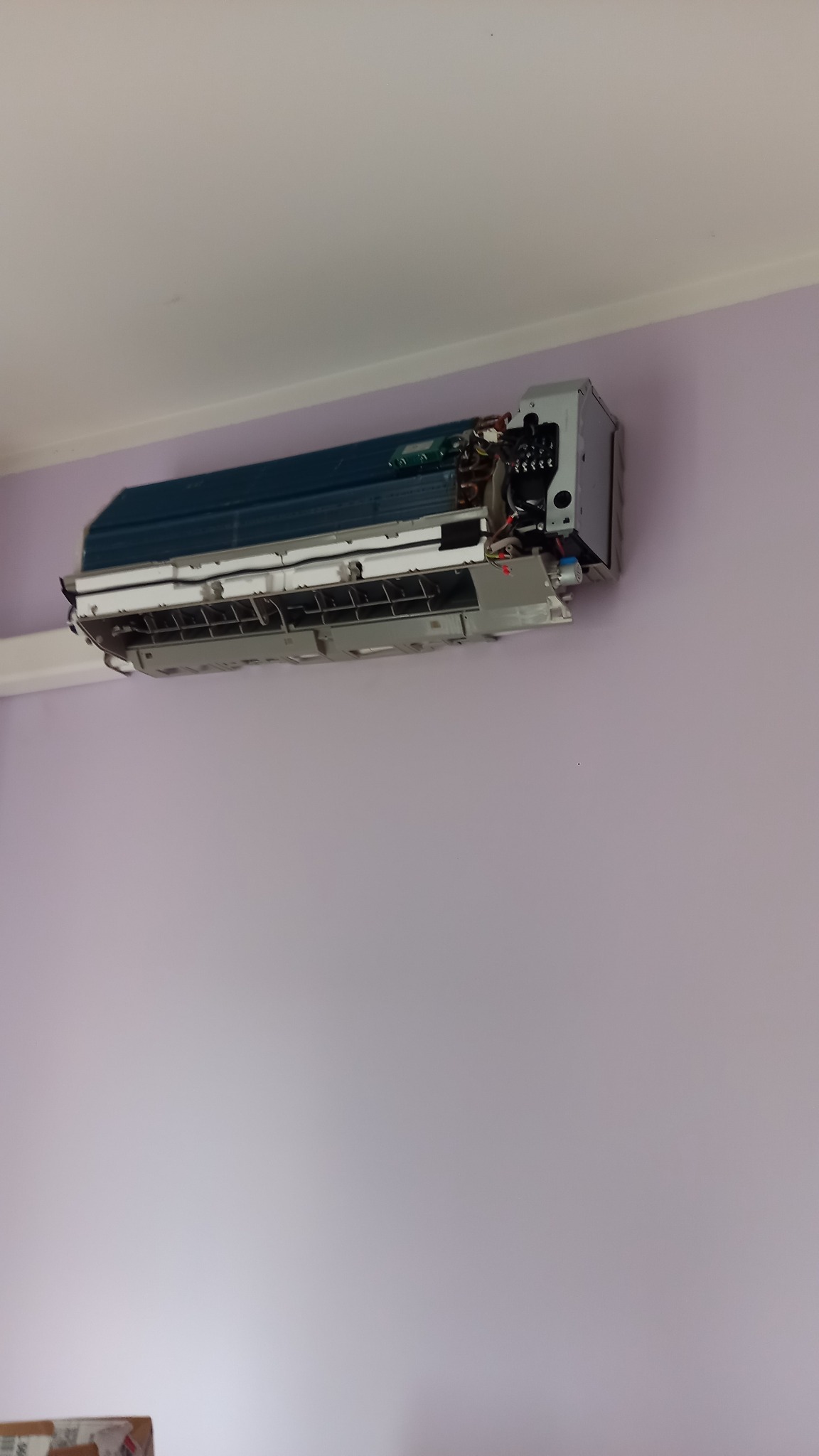 Ścienna jednostka klimatyzacji z otwartą obudową, widoczne wewnętrzne komponenty i instalacja elektryczna na tle fioletowej ściany.