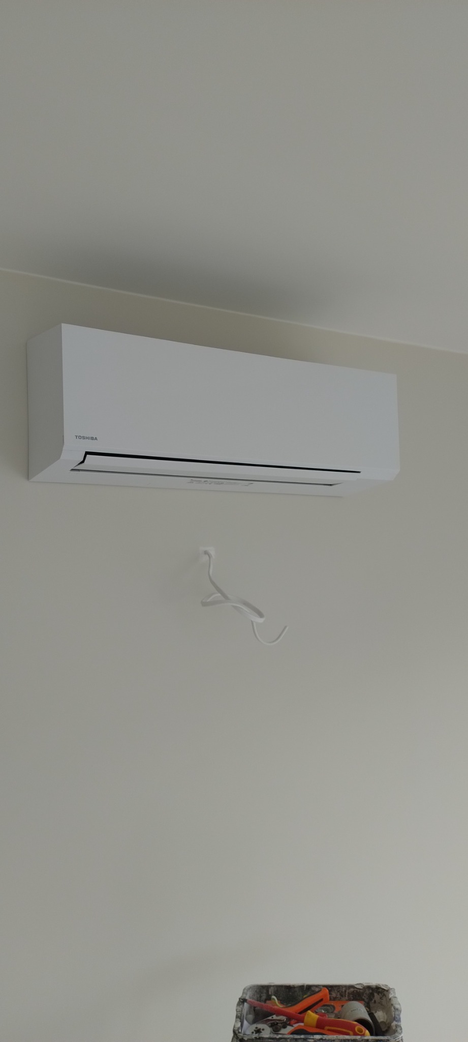 Zamontowany, biały klimatyzator Toshiba na ścianie w jasnym pomieszczeniu, z widocznymi kablami i pojemnikiem z narzędziami poniżej, co sugeruje trwający montaż.