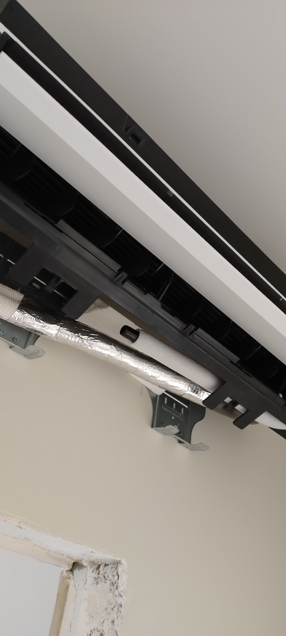 Wewnętrzna jednostka klimatyzacji z widocznymi rurami w izolacji aluminiowej, zamontowana na ścianie z niedokończonym wykończeniem w rogu pomieszczenia.