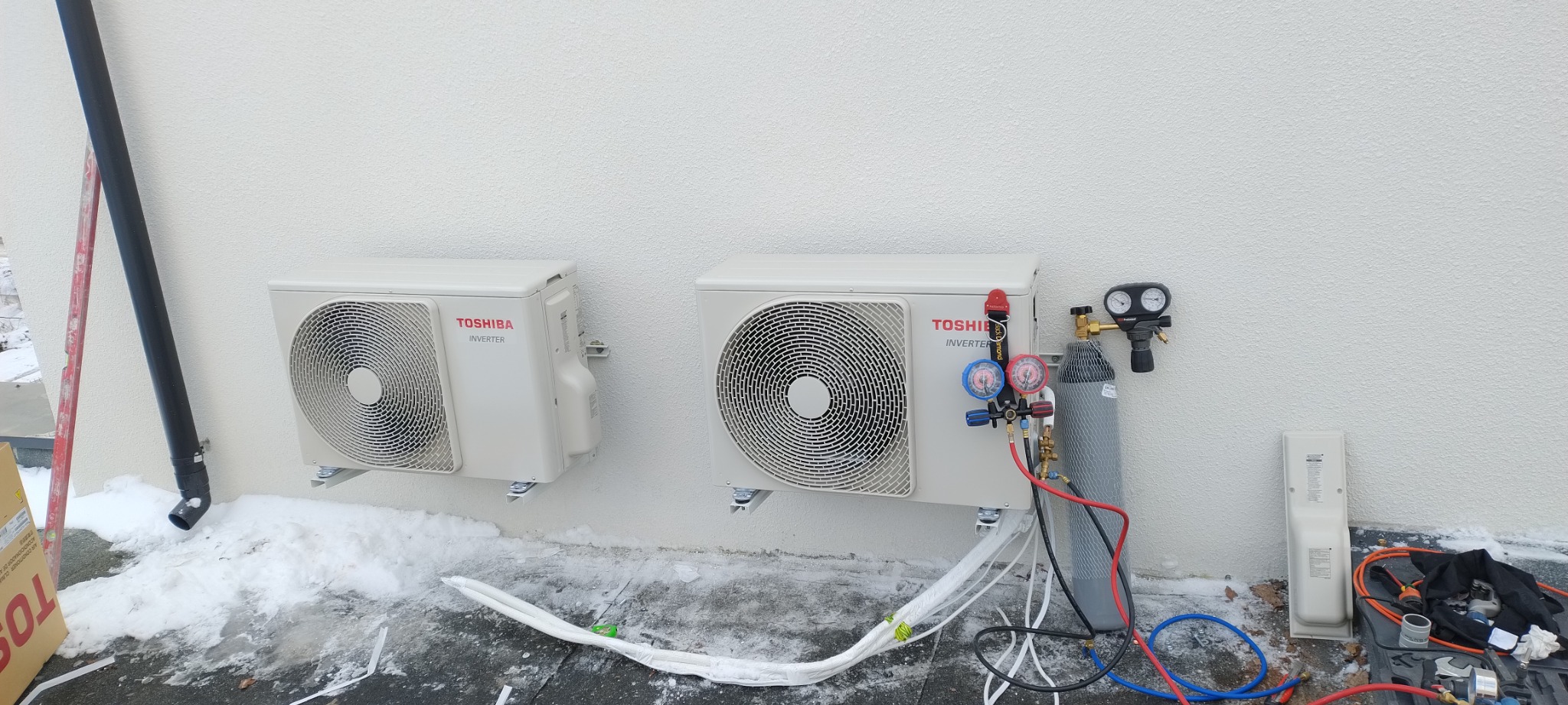 Dwa białe klimatyzatory Toshiba Inverter zamontowane na zewnątrz budynku, obok jeden z nich podłączony do butli z gazem za pomocą manometrów i przewodów, na ziemi widoczny śnieg.