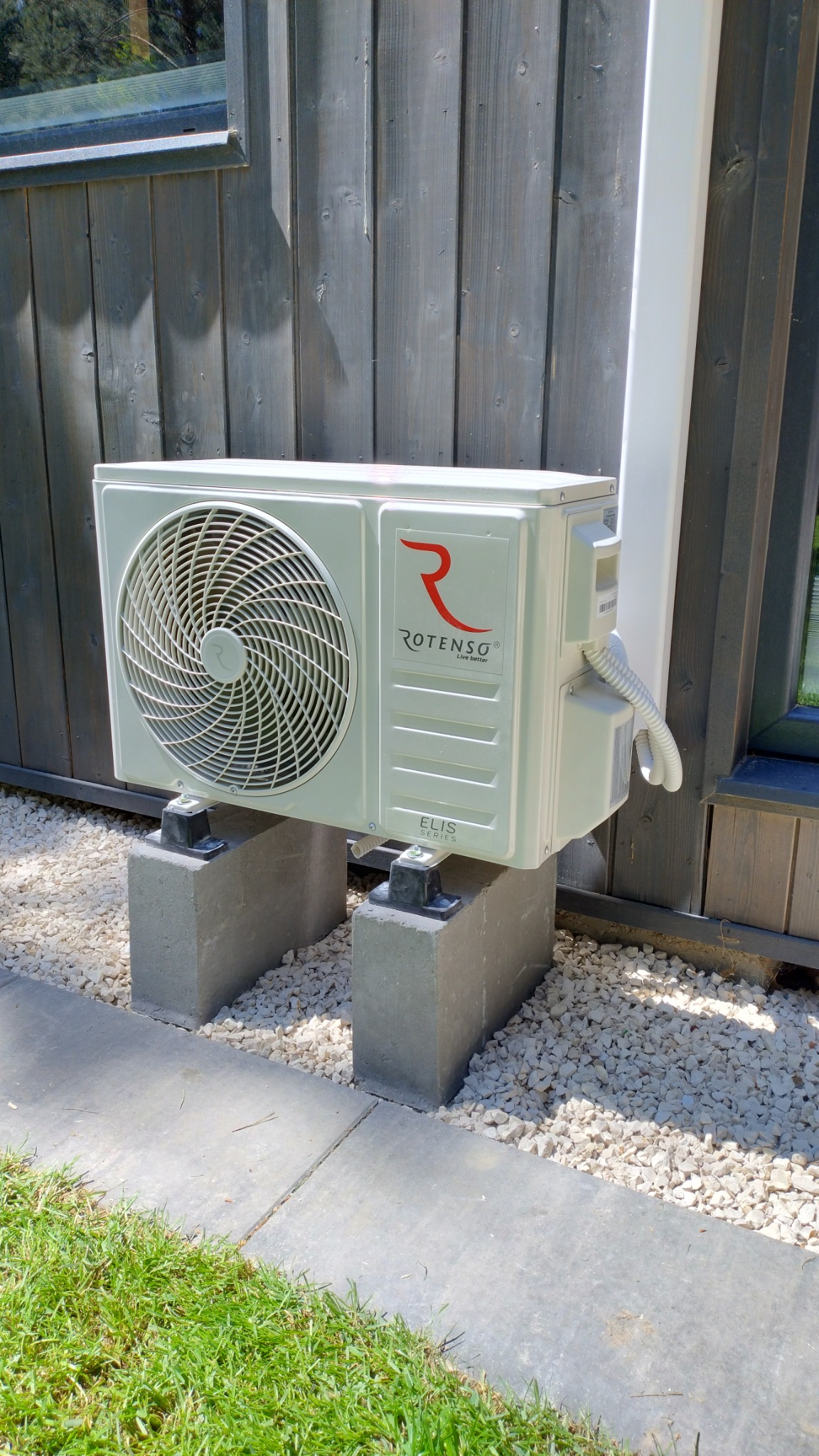 Zewnętrzna jednostka klimatyzacji Rotenso ELIS zamontowana na betonowych bloczkach przy drewnianej elewacji domu, otoczona białym żwirem.