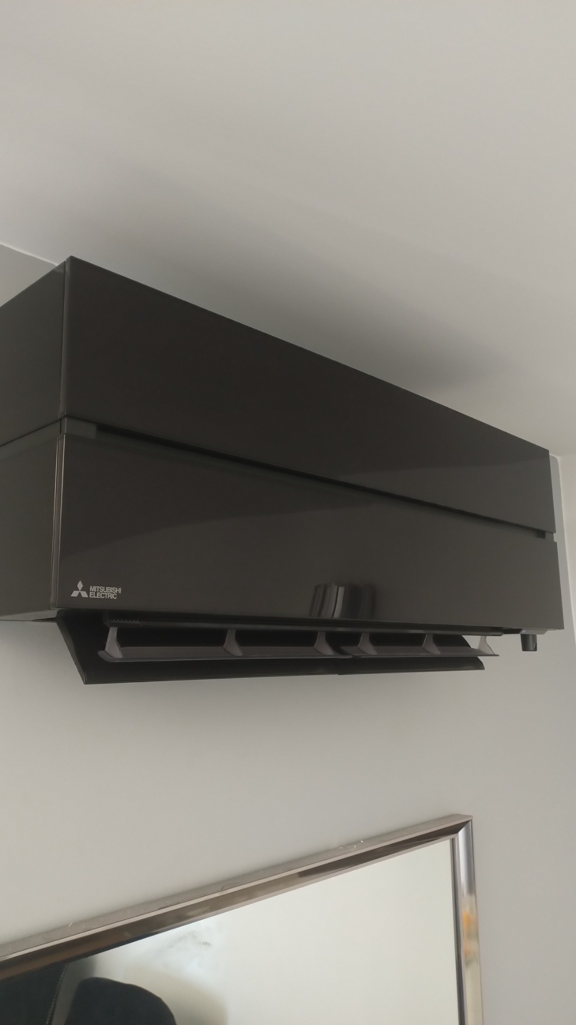 Czarna, błyszcząca klimatyzacja Mitsubishi Electric zamontowana na ścianie, widoczny fragment ramy lustra w dolnej części kadru.