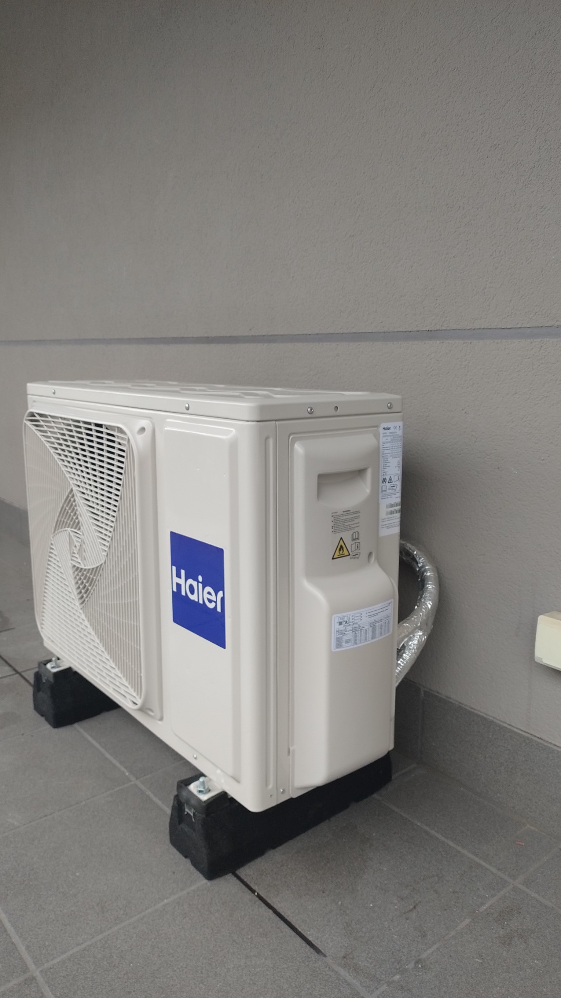 Zewnętrzna jednostka klimatyzacji Haier umieszczona na czarnych gumowych podkładkach na tle szarej ściany budynku.
