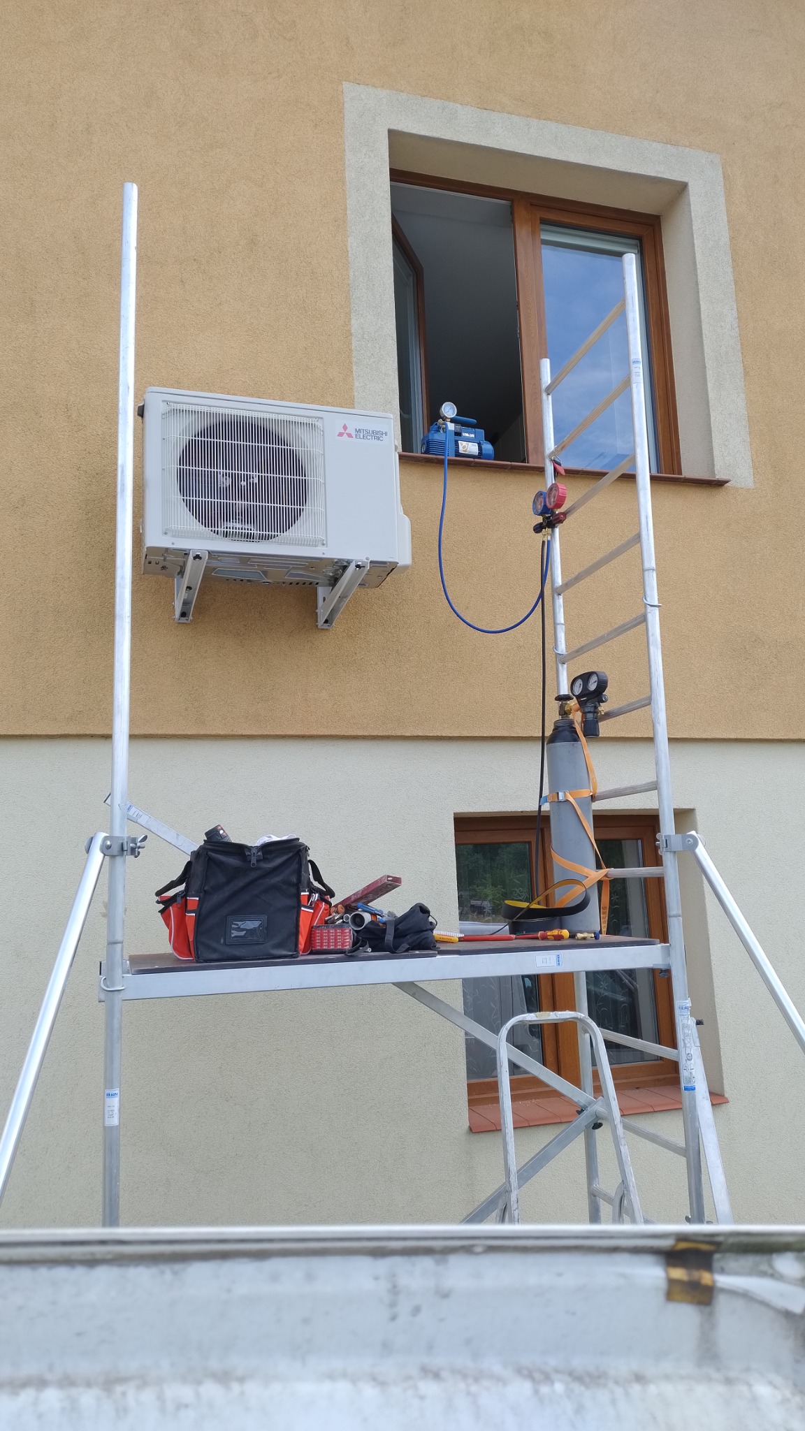 Montaż klimatyzatora Mitsubishi na elewacji domu z użyciem rusztowania; widoczna butla z gazem i pompa próżniowa w oknie.