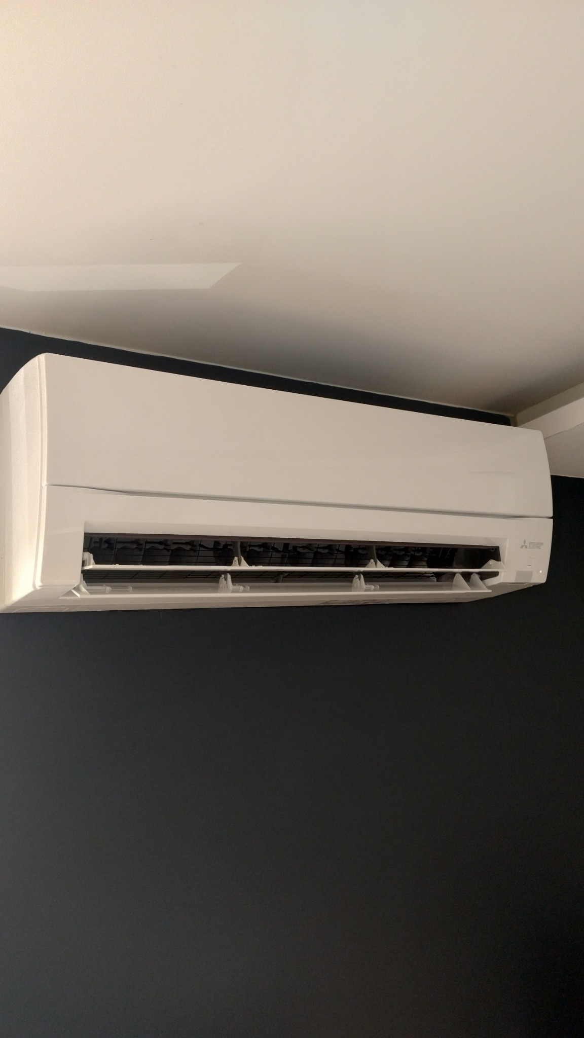 Biała jednostka klimatyzacji Mitsubishi Electric zamontowana na ciemnej ścianie w domu w Józefowie.