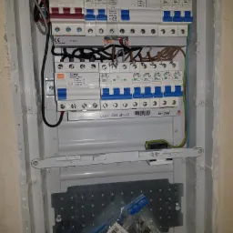 Rozdzielnica elektryczna w trakcie montażu, widoczne zabezpieczenia nadprądowe, wyłączniki różnicowoprądowe, przewody oraz akcesoria montażowe w obudowie podtynkowej.