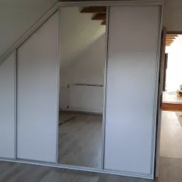 Biała szafa wnękowa z przesuwnymi drzwiami i lustrem, zamontowana na poddaszu ze skośnym sufitem, widoczna z boku otwarte drzwi do innego pomieszczenia.