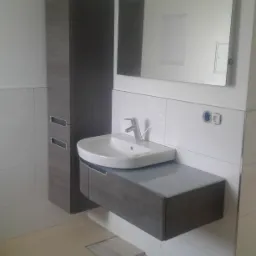 Nowoczesna łazienka z wiszącą szafką pod umywalkę w odcieniach szarości, wysoką szafką boczną z szufladami i podświetlanym lustrem, widoczne gniazdka elektryczne na białych płytkach, niedokończona...