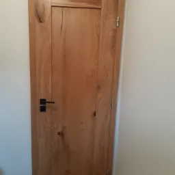 Jasne, drewniane drzwi wewnętrzne z czarną klamką i widocznymi słojami drewna, zawieszone na srebrnych zawiasach, na tle białej ściany.