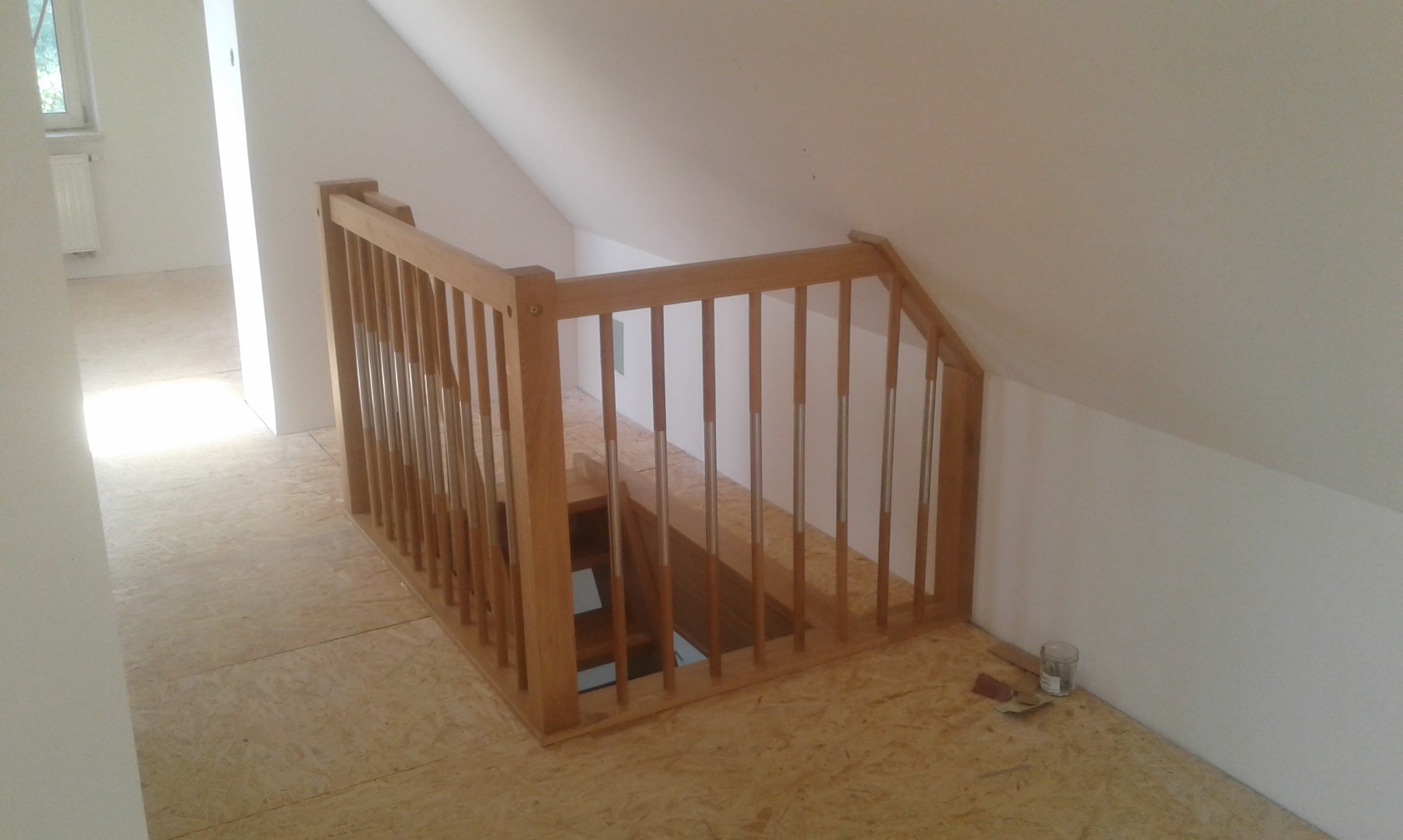 Fragment drewnianych schodów z balustradą z metalowymi prętami na poddaszu, widoczny otwór w podłodze, w tle surowe, białe ściany i podłoga z płyt OSB, obok puszka i papier ścierny.