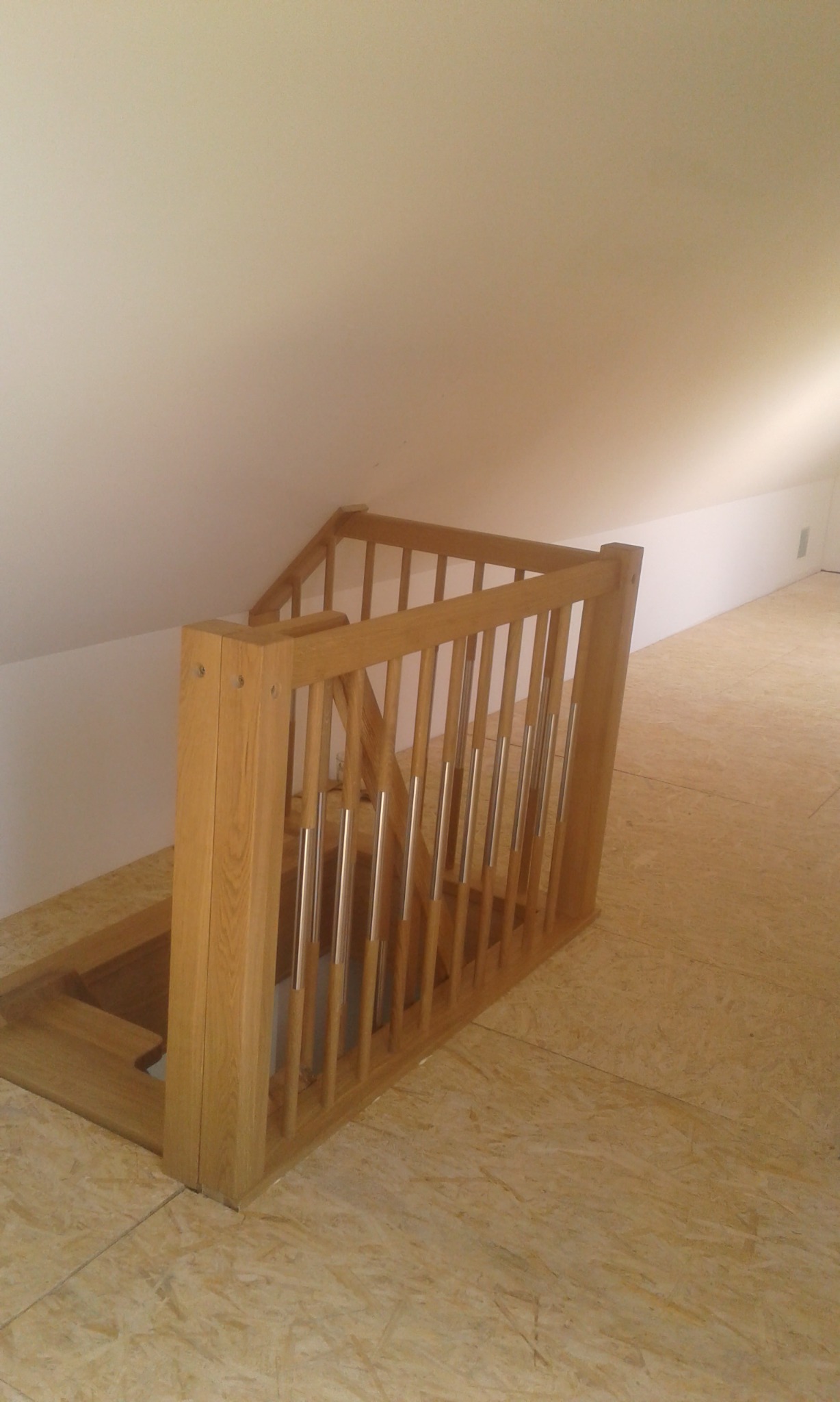 Schody strychowe z drewnianą balustradą i metalowymi tralkami w jasnym pomieszczeniu na poddaszu, widok z góry na otwór w podłodze.