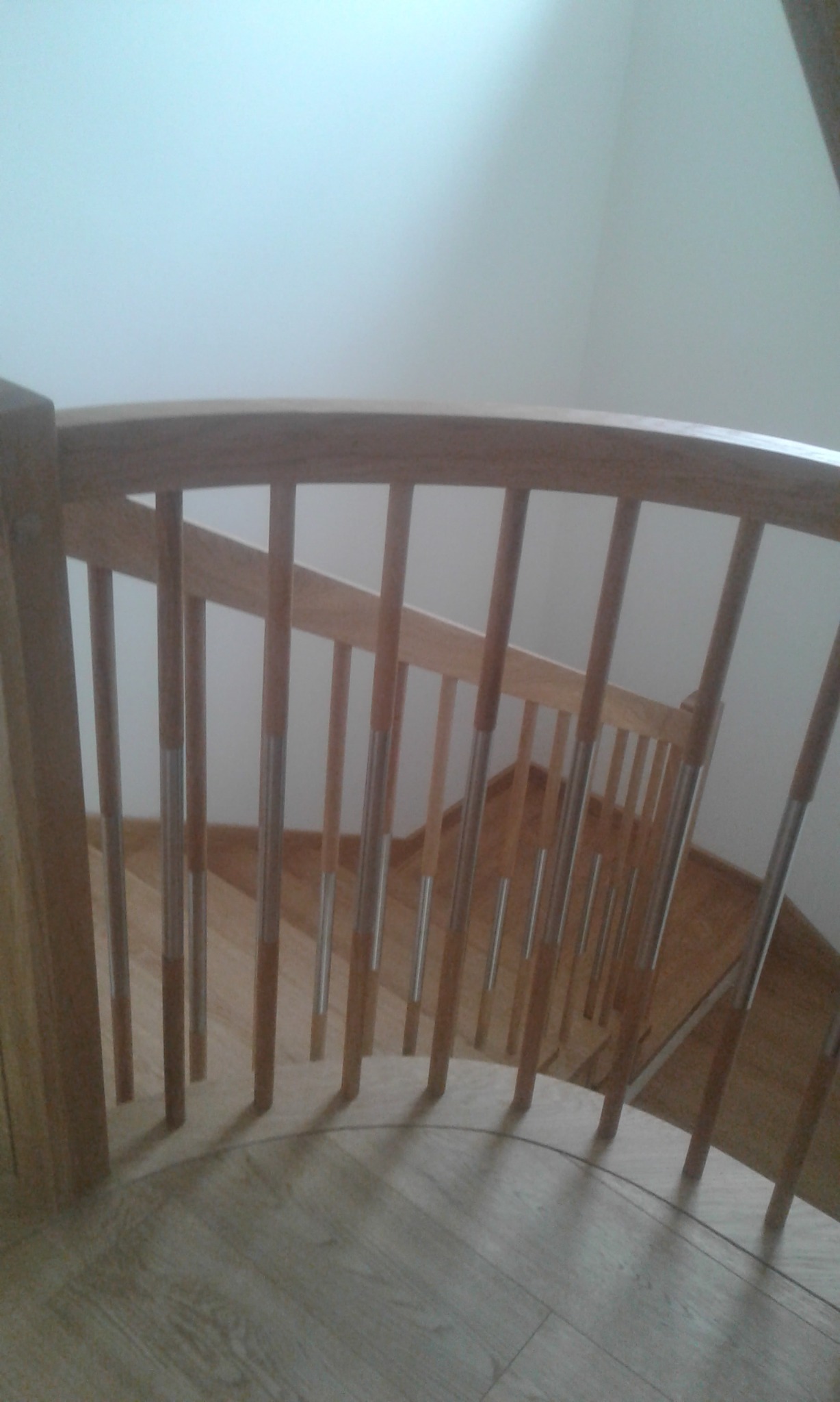 Fragment drewnianych schodów z metalowymi akcentami na poręczy, widok z góry na zakręt schodów z parkietem w tle.