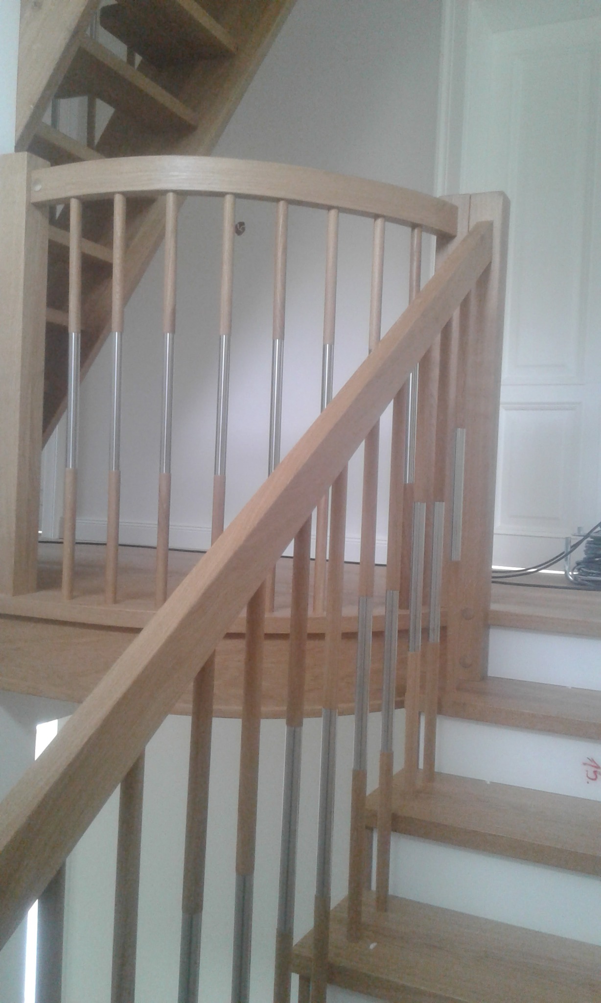 Jasne, drewniane schody z poręczą z pionowymi, metalowymi elementami, widoczne fragmenty kolejnych stopni i drzwi w tle.