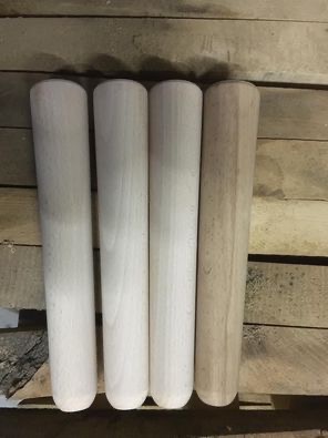 Cztery surowe, drewniane elementy o cylindrycznym kształcie, trzy jasne i jeden w odcieniu naturalnego drewna, ułożone pionowo na tle drewnianej powierzchni.