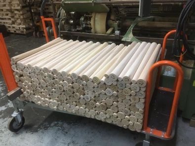 Wózek transportowy wypełniony świeżo obrobionymi, okrągłymi, drewnianymi elementami o jasnym kolorze, widoczne ślady obróbki, w tle maszyny stolarskie.