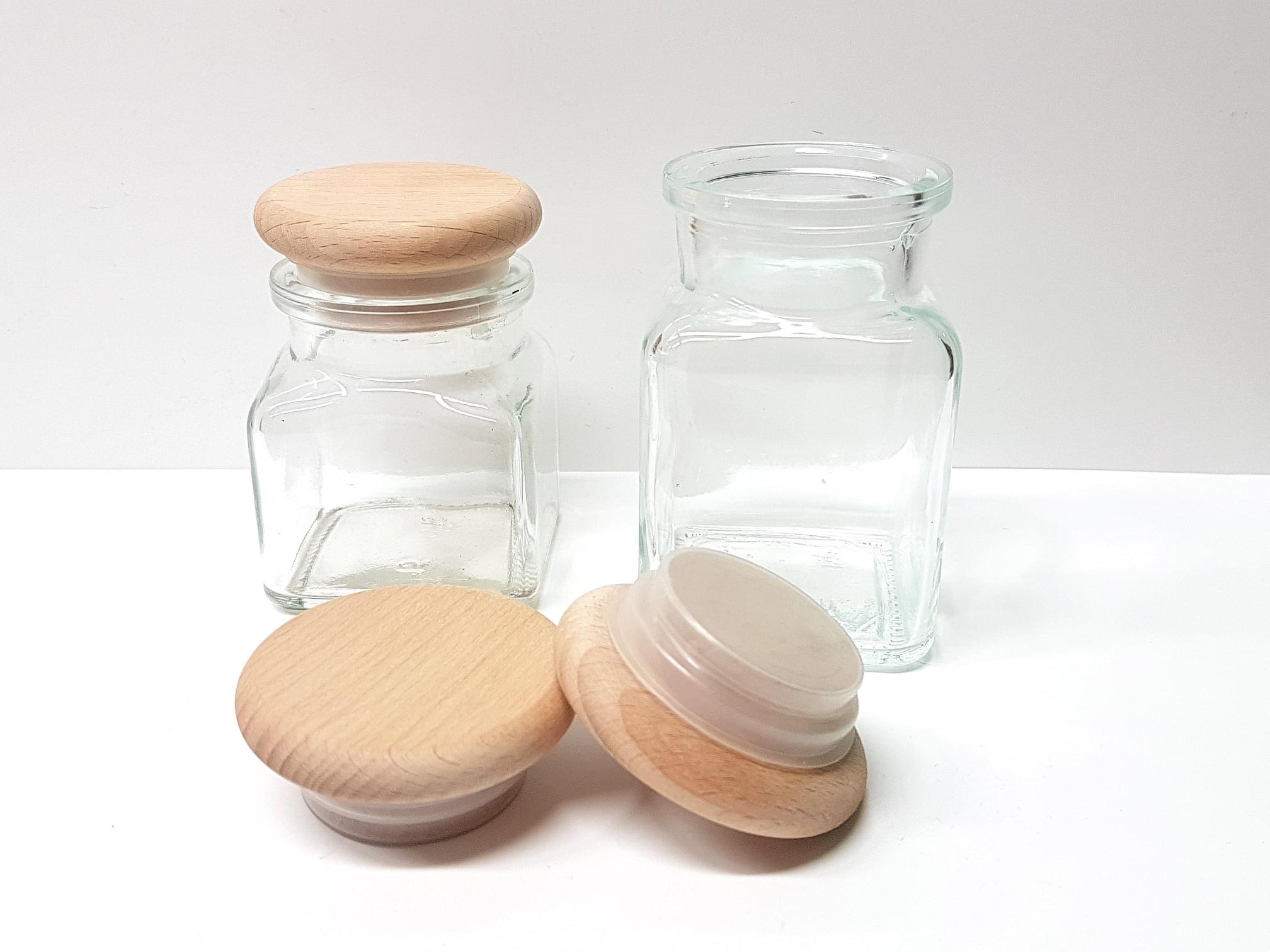 Dwa kwadratowe słoiki z przezroczystego szkła, jeden z drewnianą pokrywką, drugi otwarty, oraz dwie oddzielne drewniane pokrywki na białym tle.