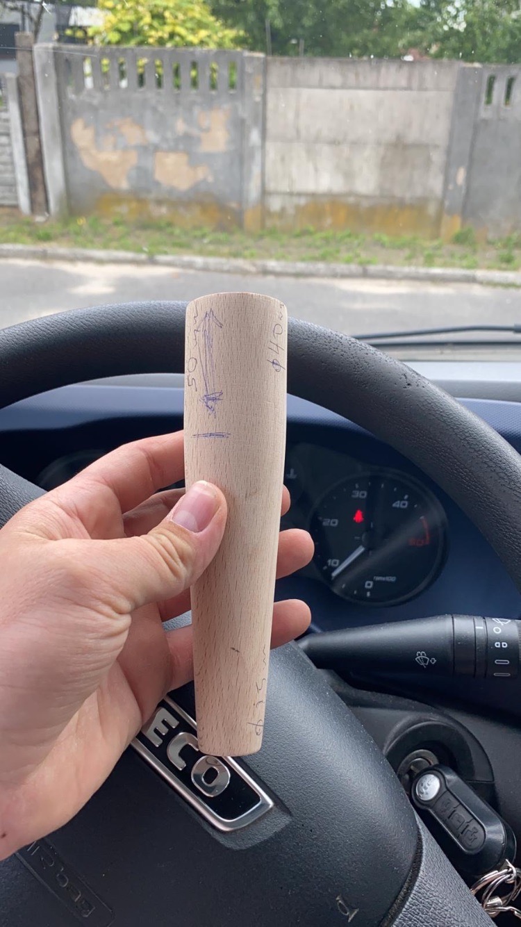 Drewniany element z odręcznymi oznaczeniami wymiarów trzymany w dłoni na tle kierownicy samochodu.