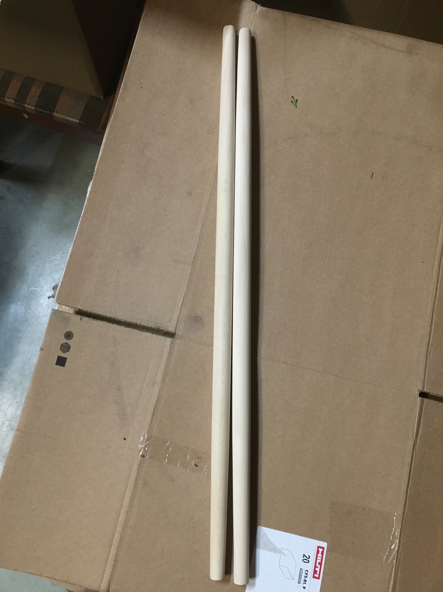 Dwa surowe, drewniane drążki leżące na kartonowym pudełku, prawdopodobnie przygotowane do dalszej obróbki.