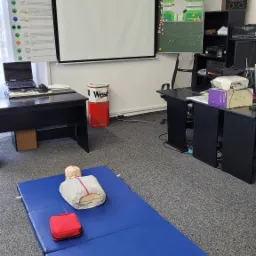 Sala szkoleniowa z manekinem do resuscytacji na niebieskiej macie, czerwona apteczka obok, w tle tablica z zasadami ruchu drogowego i rzutnik.