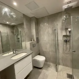 Nowoczesna łazienka z szarą płytką imitującą beton, kabina prysznicowa typu walk-in z deszczownicą, podwieszana toaleta i umywalka nablatowa na białej szafce z szarym blatem, duże lustro.