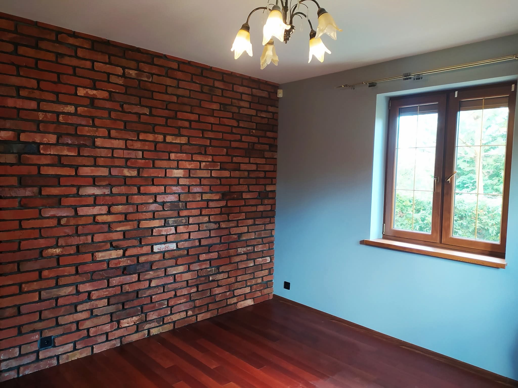 Pokój po remoncie: ceglana ściana z widoczną fakturą, drewniana podłoga i okno z drewnianą ramą. Na suficie wisi ozdobny żyrandol.