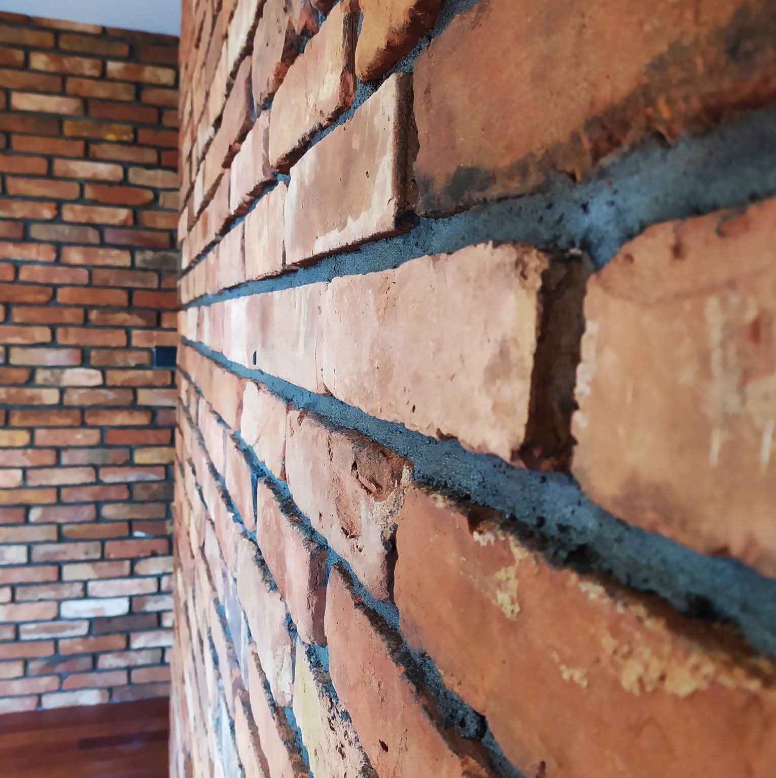 Dekoracyjna ściana z czerwonej cegły, z widoczną fakturą i szarą fugą, ułożona w pomieszczeniu z drewnianą podłogą i fragmentem drugiej ściany w tle.